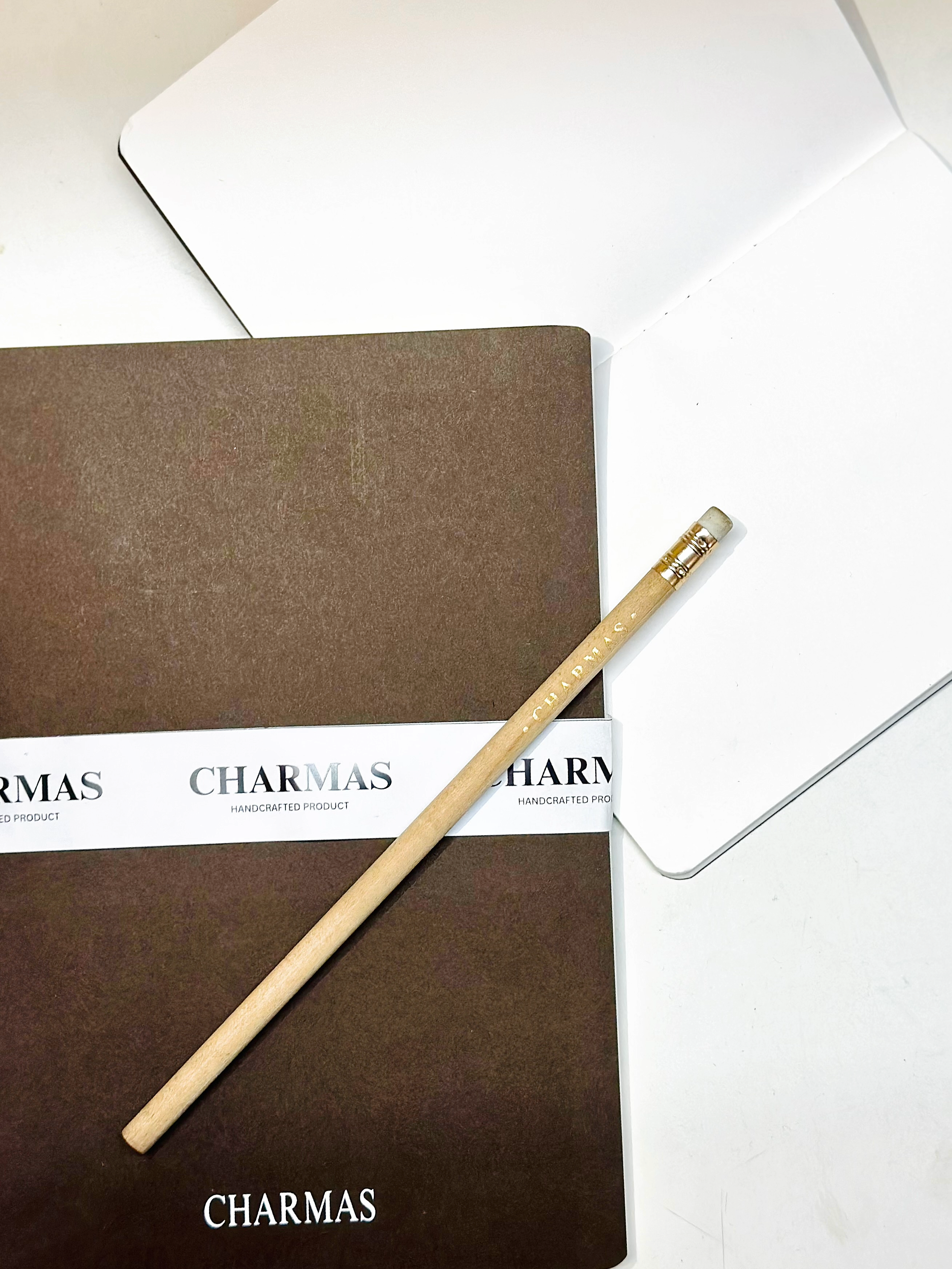 Brown notebook with a pencil on top, branded 'Charmas', against a white background