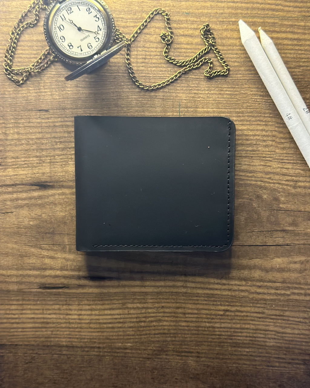 Black wallet on a wooden surface with a pocket watch and pencil.